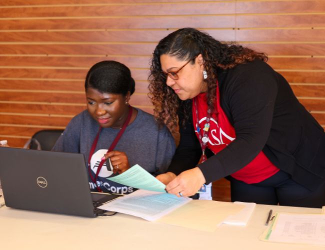 Two women, one wearing a gray T-shirt and one wearing a red T-shirt, working at a computer.